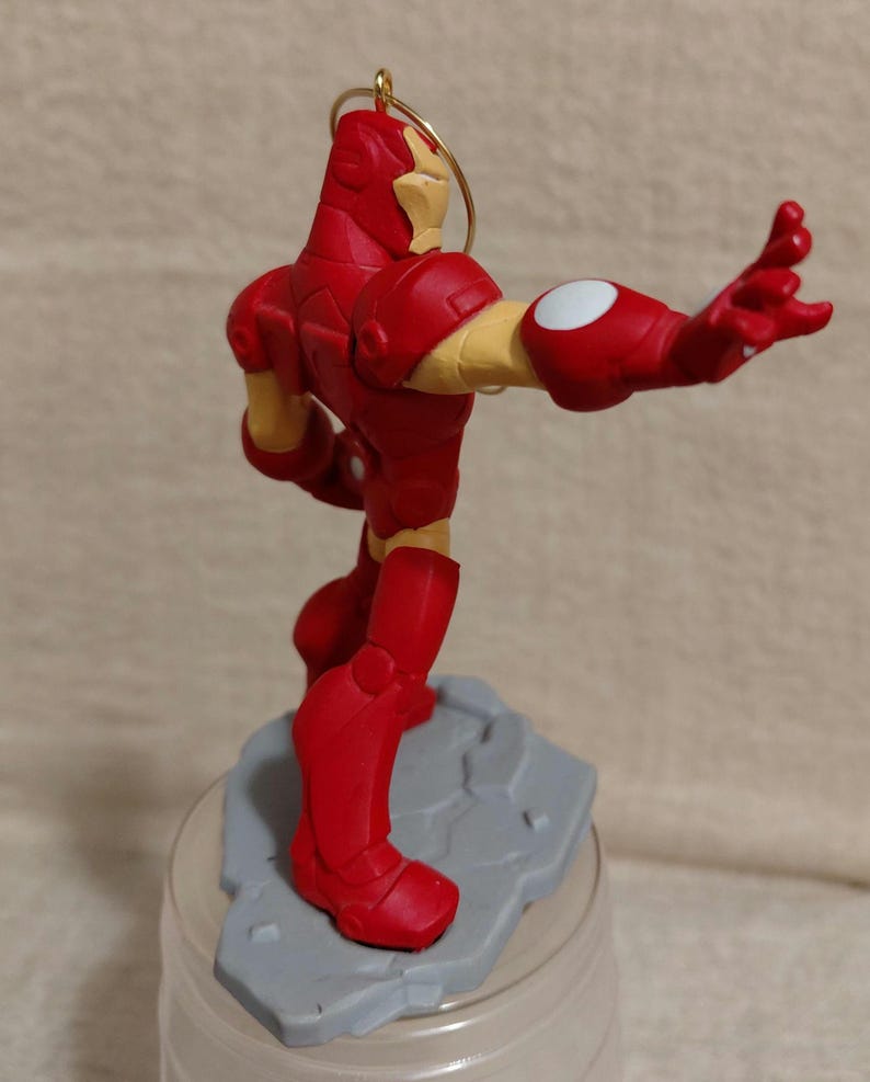 May include: A red and gold Iron Man figurine, posed with one arm extended. The figurine is on a gray base and has a gold loop for hanging. The figure's arm has a white circle on the elbow. The figurine is a collectible item.