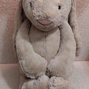 Puede incluir: Un peluche de conejo marrón claro con orejas largas y caídas. El conejo tiene una nariz rosa y ojos negros pequeños. Sus brazos y piernas están ligeramente doblados y parece estar sentado erguido. El pelaje es suave y esponjoso.