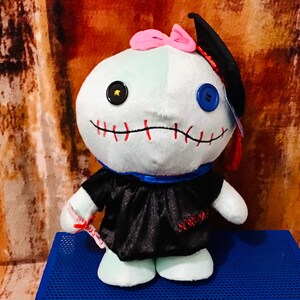 May include: A blue and white plush toy wearing a black graduation gown and cap. The toy has a stitched mouth and mismatched buttons for eyes. The toy is holding a red diploma.