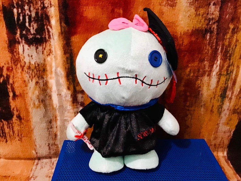 May include: A blue and white plush toy wearing a black graduation gown and cap. The toy has a stitched mouth and mismatched buttons for eyes. The toy is holding a red diploma.