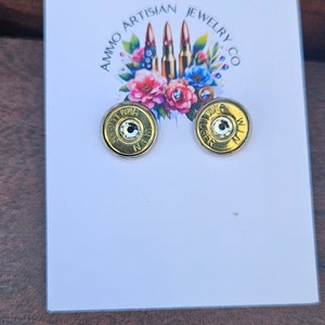 May include: A pair of gold-colored stud earrings with a clear crystal center, set on a white card with the text "Ammo Artisan Jewelry Co." and a floral design. The earrings are on a brown wooden surface.
