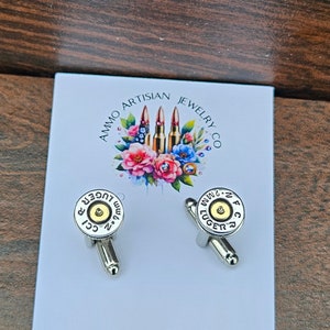 May include: Silver cufflinks with a bullet casing design. Each cufflink features a gold-colored center and the text "CCI 9mm Luger". The cufflinks are displayed on a white card with the text "Ammo Artisan Jewelry Co."