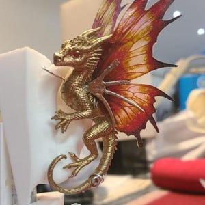 May include: A golden dragon figurine with detailed scales and vibrant red and orange wings. The dragon is posed on a white structure, showcasing its intricate design. The wings have a gradient effect, adding depth to the fantasy creature.