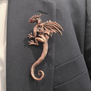 May include: A copper-toned dragon brooch featuring a textured, scaled body, folded wings, and a long, curled tail. The dragon has a small, bright red stone set as an eye. The brooch is pinned to the lapel of a dark charcoal gray suit jacket.