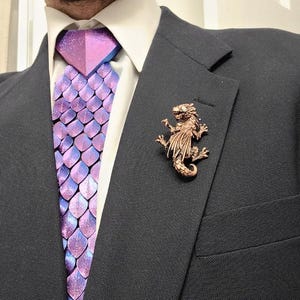 May include: A dark charcoal suit jacket worn with a white collared shirt, a purple and blue iridescent scale-patterned tie, and a metallic bronze-colored dragon-shaped lapel pin attached to the left side of the suit jacket lapel.