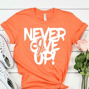 May include: Orange t-shirt with white text that reads "NEVER GIVE UP!" with a white ribbon symbol. The shirt is on a white wood background with white sneakers and pink roses.