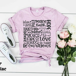 May include: A heather prism lilac t-shirt with a black graphic print of words including "believe", "peace", "faith", "hope", "trust", "you", "matter", "humble", "brave", "pray", "love", "be courageous", and "be happy".