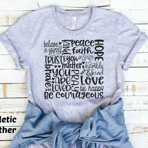 May include: A heather grey t-shirt with a black graphic design featuring the words "believe in yourself", "peace", "faith", "hope", "trust", "you inspire", "matter", "humble", "you are", "love", "be happy", and "be courageous".