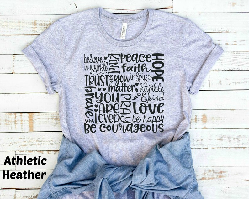 May include: A heather grey t-shirt with a black graphic design featuring the words "believe in yourself", "peace", "faith", "hope", "trust", "you inspire", "matter", "humble", "you are", "love", "be happy", and "be courageous".
