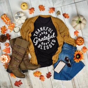 Może przedstawiać: Czarny t-shirt z napisem "Thankful Grateful Blessed" w białym k&oacute;łku. Koszulka jest stylizowana z brązowym kardiganem, niebieskimi dżinsami, brązowymi butami i jesiennymi dekoracjami.