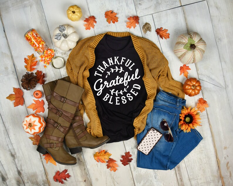 Może przedstawiać: Czarny t-shirt z napisem "Thankful Grateful Blessed" w białym k&oacute;łku. Koszulka jest stylizowana z brązowym kardiganem, niebieskimi dżinsami, brązowymi butami i jesiennymi dekoracjami.