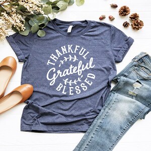 Może przedstawiać: Koszulka w kolorze granatowym z białym kołem z napisem "Thankful Grateful Blessed" i wzorem liści.