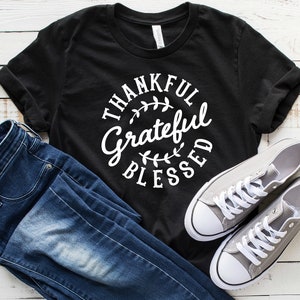 Może przedstawiać: Czarny T-shirt z białym tekstem w k&oacute;łku, kt&oacute;ry brzmi "Thankful Grateful Blessed" z małymi wzorami liści.