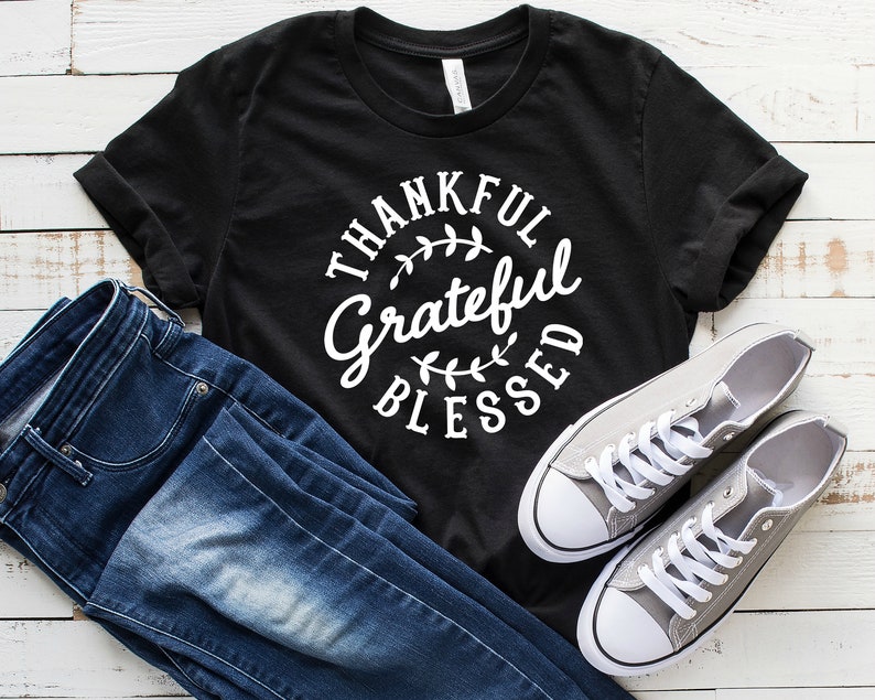 Może przedstawiać: Czarny T-shirt z białym tekstem w k&oacute;łku, kt&oacute;ry brzmi "Thankful Grateful Blessed" z małymi wzorami liści.