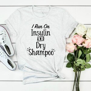 Puede incluir: Una camiseta gris con el texto "I Run On Insulin And Dry Shampoo" impreso en ella. La camiseta est&aacute; colocada sobre una superficie de madera blanca con un par de zapatillas blancas y un ramo de rosas rosas y blancas.