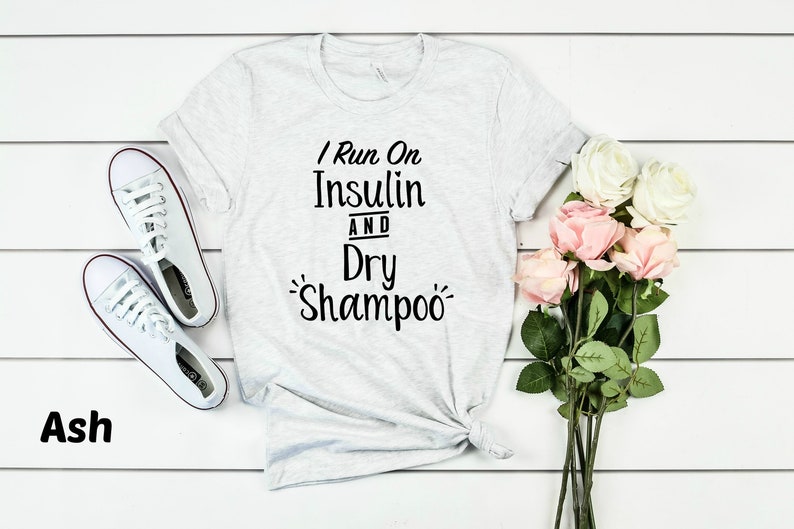 Puede incluir: Una camiseta gris con el texto "I Run On Insulin And Dry Shampoo" impreso en ella. La camiseta est&aacute; colocada sobre una superficie de madera blanca con un par de zapatillas blancas y un ramo de rosas rosas y blancas.