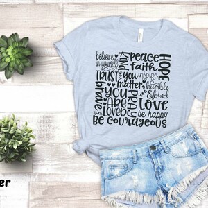 May include: A light blue t-shirt with a black word art design that reads "Believe in yourself, Peace, Faith, Trust you, Inspire, Matter, Humble, You are loved, Be happy, Be courageous".