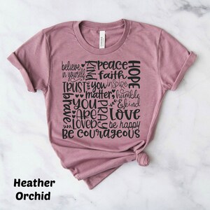May include: A heather orchid colored t-shirt with a graphic design of words in black. The words include "believe", "peace", "faith", "hope", "trust", "you", "matter", "humble", "brave", "are", "love", "be happy", and "be courageous".