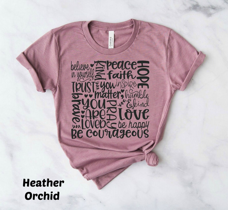 May include: A heather orchid colored t-shirt with a graphic design of words in black. The words include "believe", "peace", "faith", "hope", "trust", "you", "matter", "humble", "brave", "are", "love", "be happy", and "be courageous".