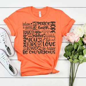 May include: Orange t-shirt with a black graphic print of words like "believe", "peace", "hope", "trust", "love", and "be courageous".