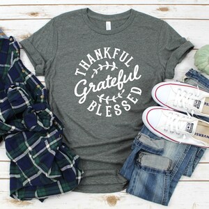 Może przedstawiać: Szary t-shirt z nadrukiem "Thankful Grateful Blessed" w białym k&oacute;łku z liściastym wzorem. Koszulka jest pokazana z koszulą w kratę, niebieskimi dżinsami i białymi butami sportowymi.