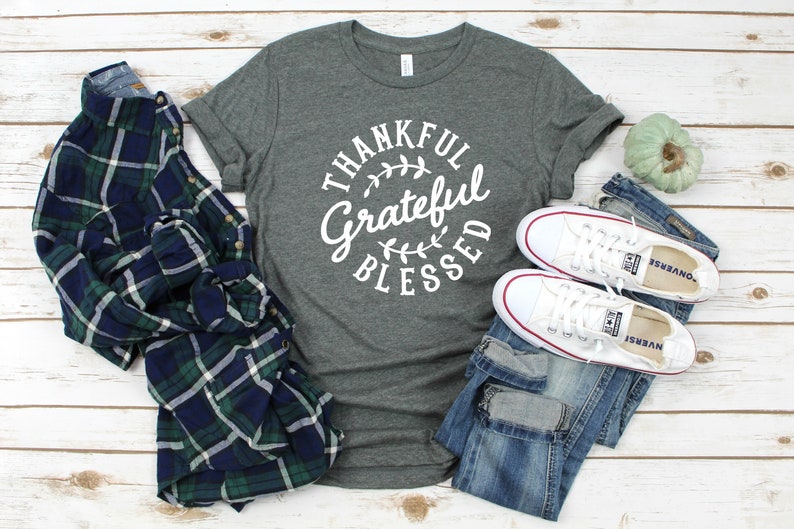Może przedstawiać: Szary t-shirt z nadrukiem "Thankful Grateful Blessed" w białym k&oacute;łku z liściastym wzorem. Koszulka jest pokazana z koszulą w kratę, niebieskimi dżinsami i białymi butami sportowymi.
