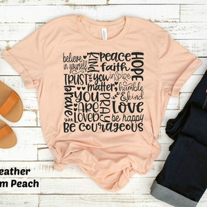 May include: A peach-colored t-shirt with a black graphic design featuring the words "believe", "peace", "hope", "trust", "you", "matter", "love", "be courageous", and more. The shirt is made of a soft, comfortable material.