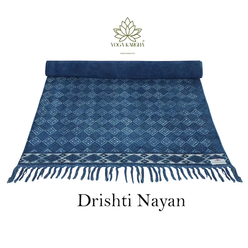 May include: A blue cotton yoga mat with a white diamond pattern and fringe. The mat is rolled up and has a label that says "Yoga Kargha" and "Abode Crafts". The text "Dristhi Nayan" is written below the mat.