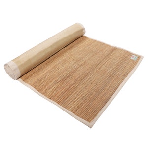 K&ouml;nnte beinhalten: Eine zusammengerollte Yogamatte aus Naturfasern mit wei&szlig;em Rand. Die Matte besteht aus gewebtem Stroh und ist f&uuml;r die Verwendung auf dem Boden konzipiert.