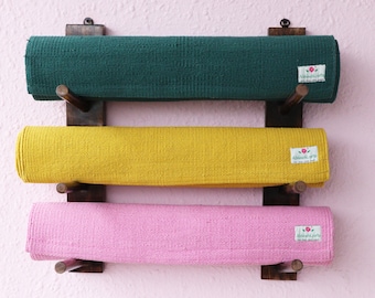 May include: Three yoga mats are rolled up and displayed on a wooden wall rack. The mats are green, yellow, and pink. Each mat has a small label with the text "Alhambra crafts" and a pink flower.