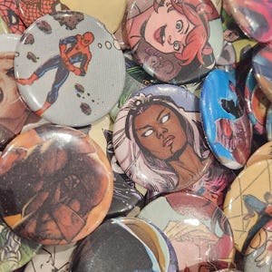 Può includere: Una collezione di spille rotonde e lucide con illustrazioni di fumetti. Le spille presentano vari personaggi, tra cui Spider-Man e Tempesta, e altri personaggi dei fumetti. Le spille sono in vari colori e design.