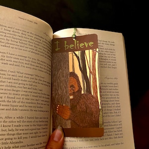 May include: A brown bookmark with the text "I believe" in green letters. The bookmark features a cartoon illustration of a brown Bigfoot standing behind a tree in a forest. The Bigfoot is smiling and has a friendly expression.
