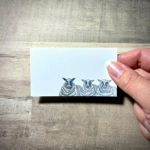 Könnte beinhalten: Eine rechteckige Karte mit einer Aquarellillustration von drei Schafen. Die Schafe sind in Grau- und Weißtönen dargestellt, mit detaillierten Wollstrukturen und dunklen Augen. Die Karte wird vor einem Holzgrund gehalten.