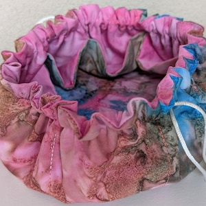 May include: A round, drawstring fabric pouch with a pink, blue, and brown marbled pattern. The pouch has a ruffled edge and a white drawstring closure. The interior fabric features a similar marbled design.