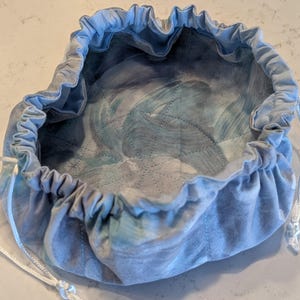 May include: A round, fabric drawstring pouch featuring a light blue and grey marbled pattern. The interior base is quilted with a geometric stitch design, and the top edge is gathered with a white cord to create a cinched opening.