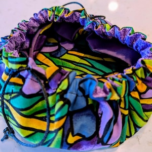 May include: A round fabric pouch with a drawstring closure, featuring a vibrant stained-glass inspired pattern in shades of purple, green, blue, and yellow. The interior is lined with solid purple fabric, and the exterior has a gathered top edge.