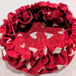 May include: A red drawstring bag with a white heart pattern. The bag's interior and exterior are red, with white hearts. The top edge is gathered with a drawstring closure. The bag is designed to be open and accessible.