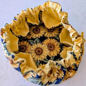 May include: A round fabric drawstring pouch featuring a dark navy blue exterior with a vibrant yellow sunflower pattern. The interior is lined with solid yellow fabric and includes multiple interior pockets along the perimeter of the bag.
