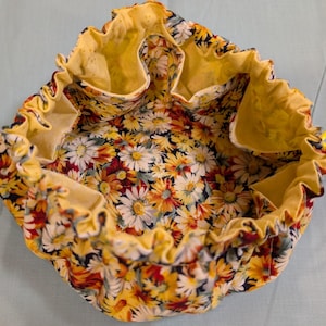 May include: A round, open drawstring bag with a floral daisy pattern. The interior and trim are yellow. The bag has several pockets and yellow drawstrings. The daisy pattern includes white, yellow, orange, and red flowers on a black background.