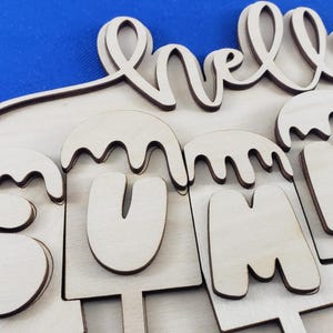 K&ouml;nnte beinhalten: Holzschild mit den Worten "hello SUMMER" ausgeschnitten. Das Wort "hello" ist kursiv geschrieben, und "SUMMER" ist in Gro&szlig;buchstaben gestaltet, die wie Eis am Stiel aussehen. Das Schild ist aus hellem Holz.