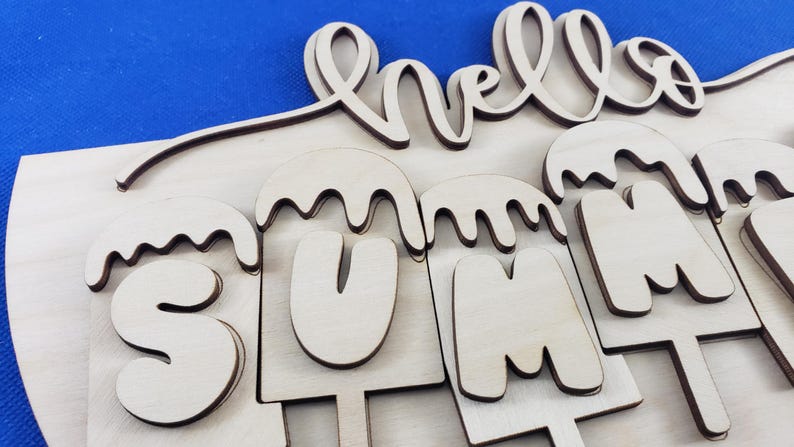K&ouml;nnte beinhalten: Holzschild mit den Worten "hello SUMMER" ausgeschnitten. Das Wort "hello" ist kursiv geschrieben, und "SUMMER" ist in Gro&szlig;buchstaben gestaltet, die wie Eis am Stiel aussehen. Das Schild ist aus hellem Holz.