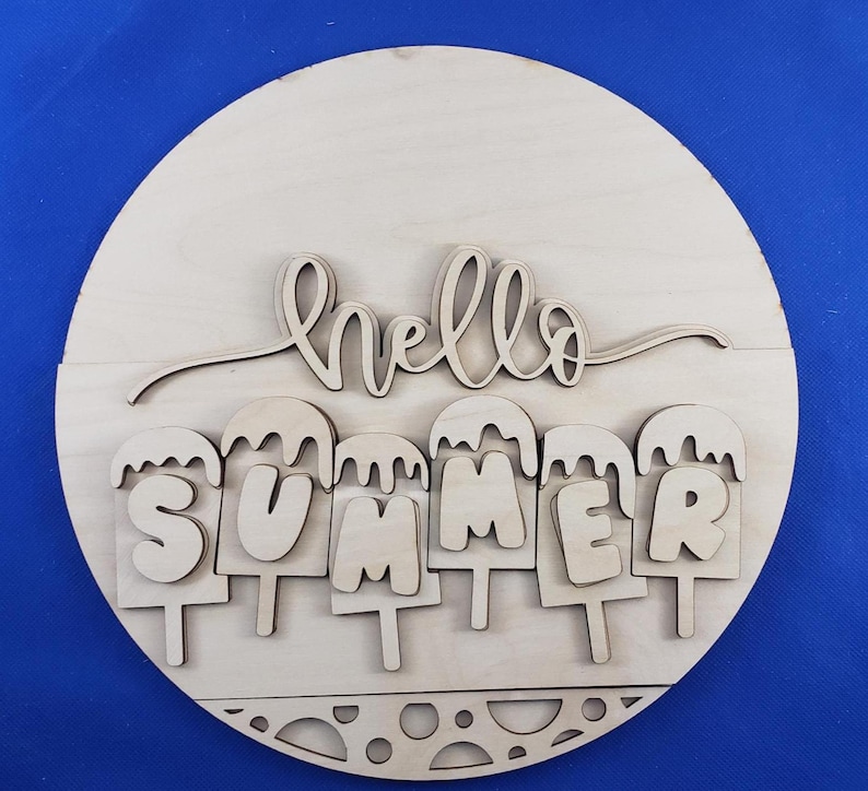 K&ouml;nnte beinhalten: Runder Holzschild mit den ausgeschnittenen Worten "hello SUMMER". Das Wort "hello" ist kursiv geschrieben, und "SUMMER" ist in Blockbuchstaben gehalten, wobei jeder Buchstabe als Eis am Stiel gestaltet ist. Das Schild ist aus unbehandeltem Holz.
