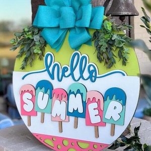 K&ouml;nnte beinhalten: Runder Holzschild mit leuchtend gr&uuml;nem und wei&szlig;em Hintergrund, auf dem die Worte "hello SUMMER" in einer verspielten Schriftart stehen. Das Schild ist mit bunten Eis am Stiel-Designs und einer gro&szlig;en t&uuml;rkisfarbenen Schleife verziert.