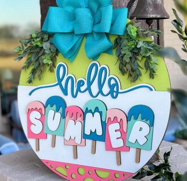 K&ouml;nnte beinhalten: Runder Holzschild mit leuchtend gr&uuml;nem und wei&szlig;em Hintergrund, auf dem die Worte "hello SUMMER" in einer verspielten Schriftart stehen. Das Schild ist mit bunten Eis am Stiel-Designs und einer gro&szlig;en t&uuml;rkisfarbenen Schleife verziert.