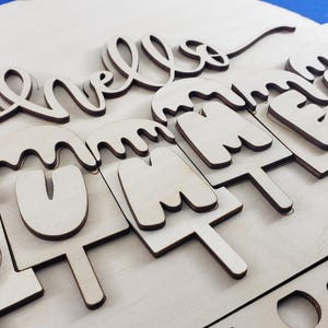 K&ouml;nnte beinhalten: Runder Holzschild mit der Aufschrift "hello SUMMER". Das Wort "hello" ist in Schreibschrift gehalten, und "SUMMER" ist als schmelzende Eis am Stiel gestaltet. Das Schild ist aus hellem Holz gefertigt.