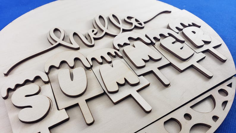 K&ouml;nnte beinhalten: Runder Holzschild mit der Aufschrift "hello SUMMER". Das Wort "hello" ist in Schreibschrift gehalten, und "SUMMER" ist als schmelzende Eis am Stiel gestaltet. Das Schild ist aus hellem Holz gefertigt.