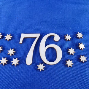 May include: Wooden number 76 with a white background and surrounded by small wooden stars on a blue surface.