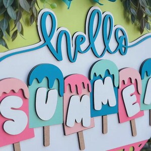 K&ouml;nnte beinhalten: Runder Holzschild mit den Worten "hello SUMMER" in T&uuml;rkis und Wei&szlig;. Das Wort "SUMMER" ist mit Buchstaben in Eisform in Rosa, T&uuml;rkis und Wei&szlig; geschrieben. Gr&uuml;ne Bl&auml;tter und ein hellgr&uuml;ner Hintergrund vervollst&auml;ndigen das Design.