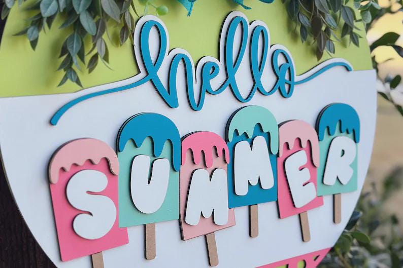 K&ouml;nnte beinhalten: Runder Holzschild mit den Worten "hello SUMMER" in T&uuml;rkis und Wei&szlig;. Das Wort "SUMMER" ist mit Buchstaben in Eisform in Rosa, T&uuml;rkis und Wei&szlig; geschrieben. Gr&uuml;ne Bl&auml;tter und ein hellgr&uuml;ner Hintergrund vervollst&auml;ndigen das Design.