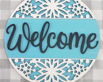 Pode incluir: Uma placa de boas-vindas redonda de madeira com um centro azul claro e um design de floco de neve branco. A palavra "Welcome" está escrita em letras azuis escuras. Perfeito para uma porta da frente ou entrada.